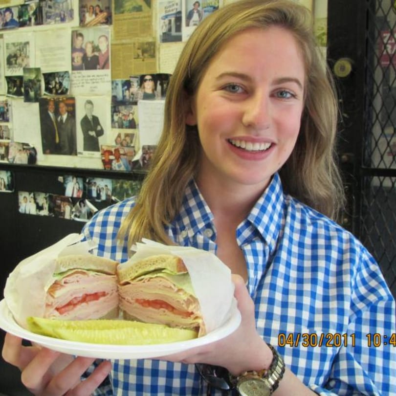 Smiling woman in a blue gingham shirt holding a large deli sandwich and pickle.