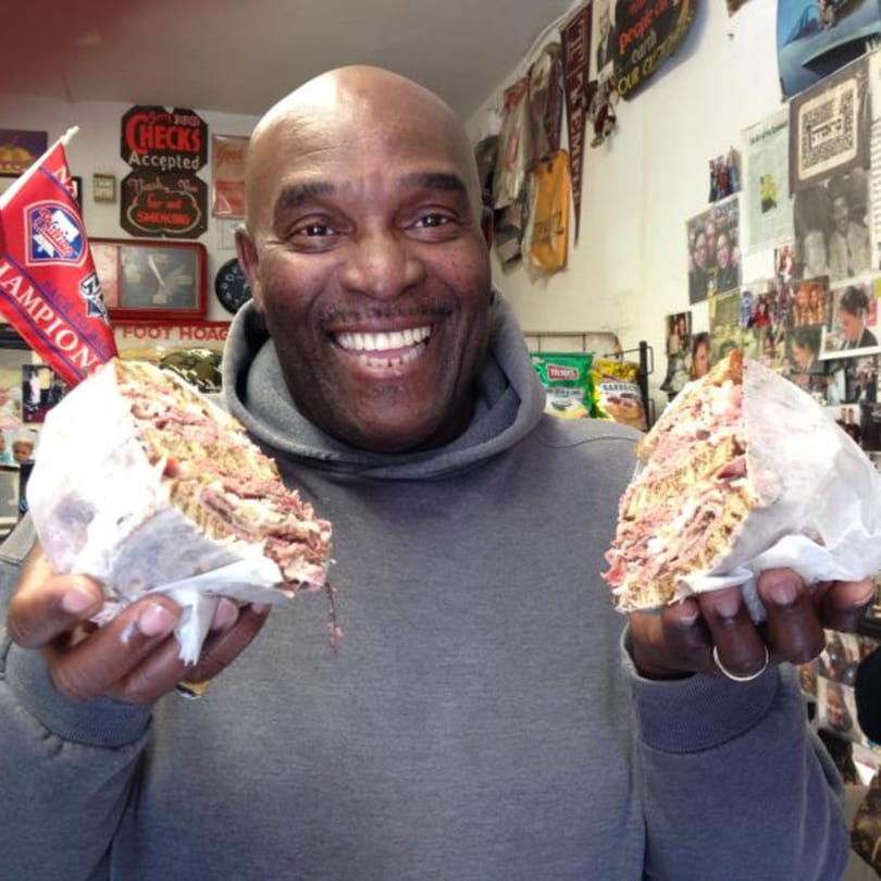 Smiling man holding two halves of a large, meat-filled deli sandwich at Koch's Deli.