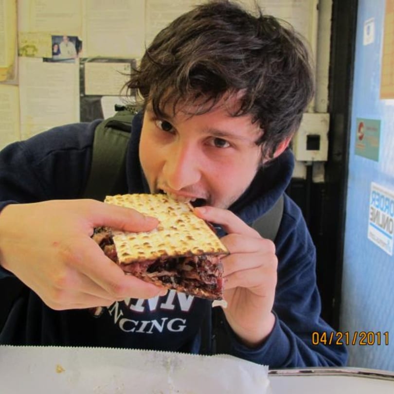 A young man in a deli eagerly takes a bite out of a large matzo sandwich.
