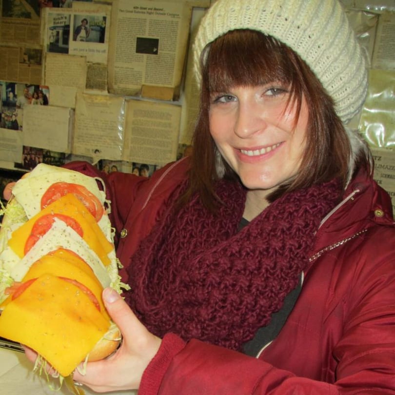Smiling woman in a white beanie and red jacket holding a large deli sandwich with cheese and tomatoes.