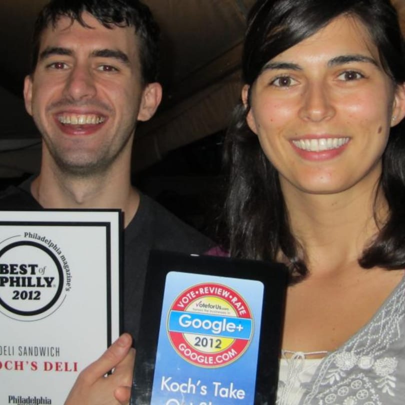 Two smiling people, a man and a woman, holding Koch's Deli awards for "Best of Philly" and Google+.