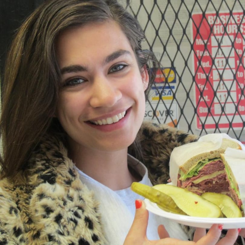 A smiling woman in a leopard print coat holds a large deli sandwich and pickles.