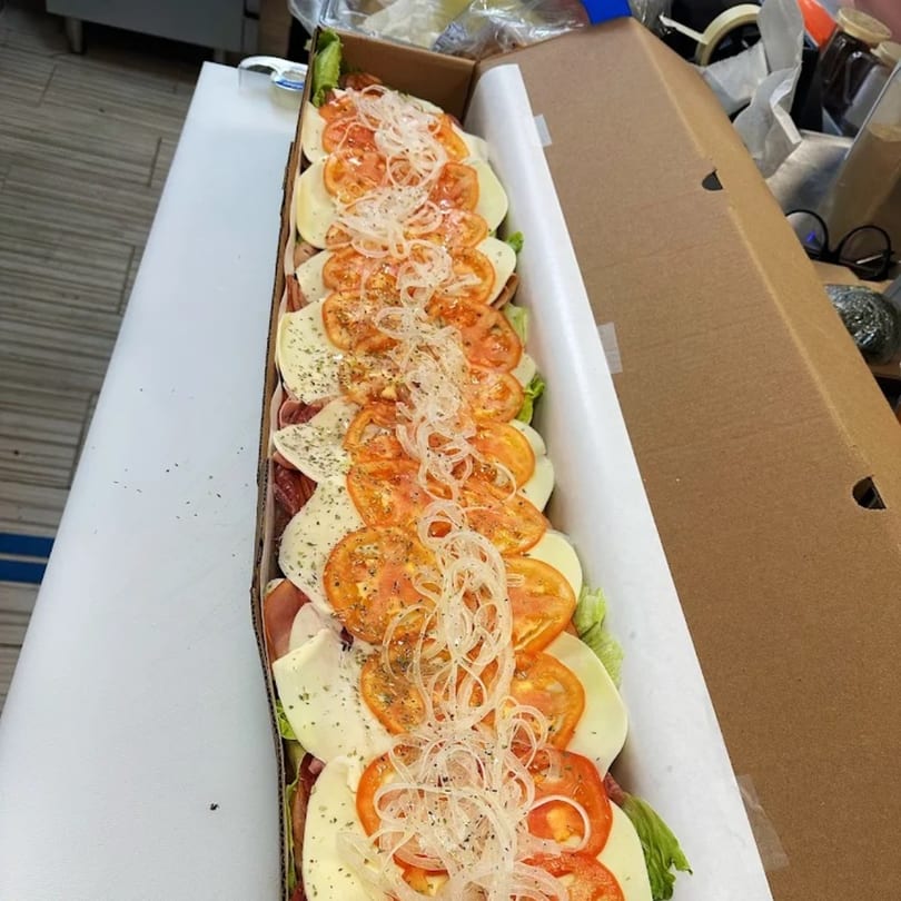 A long deli sub sandwich, topped with tomatoes, onions, and cheese, in a cardboard box.