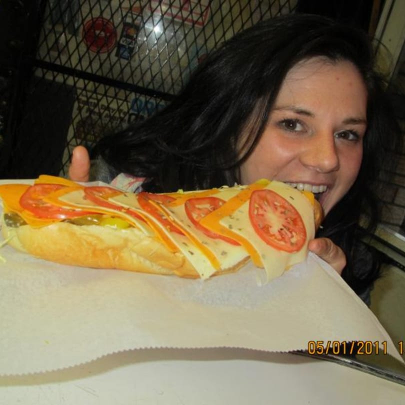 A woman smiles while taking a bite of a large deli sandwich with cheese, tomatoes, and pickles.