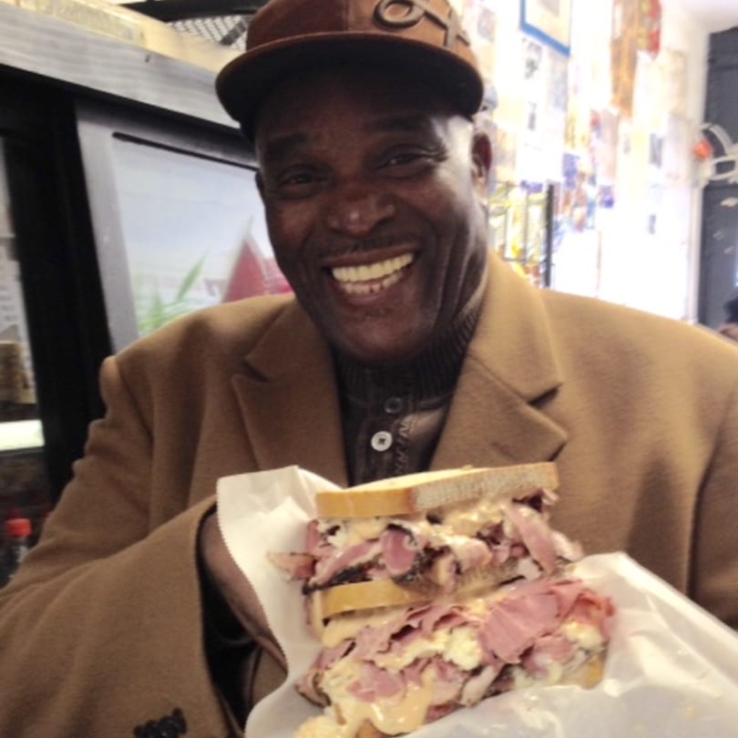 A smiling man in a brown hat and coat holds a large, overflowing deli sandwich.