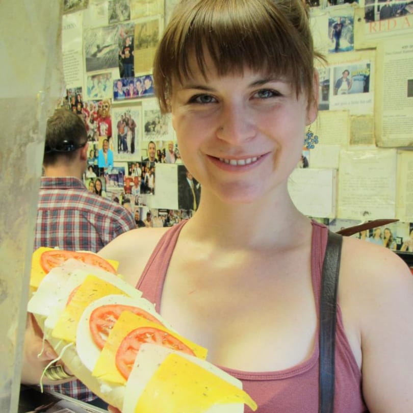 A smiling woman holds a large Koch's Deli sandwich with cheese and tomatoes.