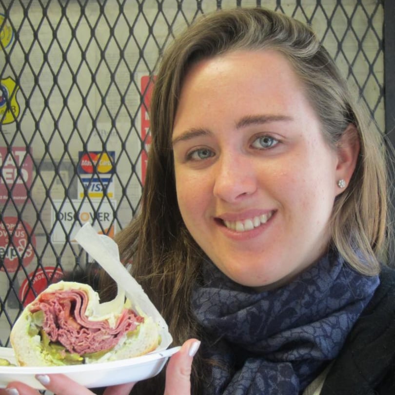 A smiling woman holds a large pastrami sandwich from Koch's Deli.