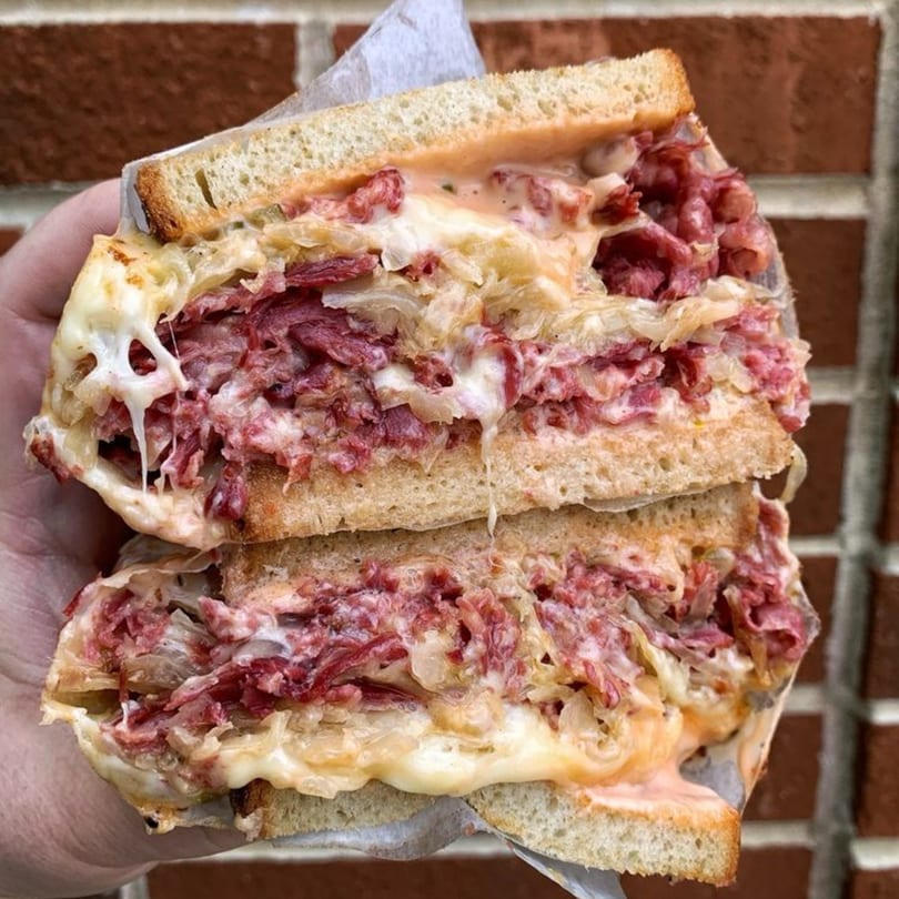 A hand holds a large, sliced Koch's Deli Reuben sandwich with corned beef, sauerkraut, cheese, and dressing.