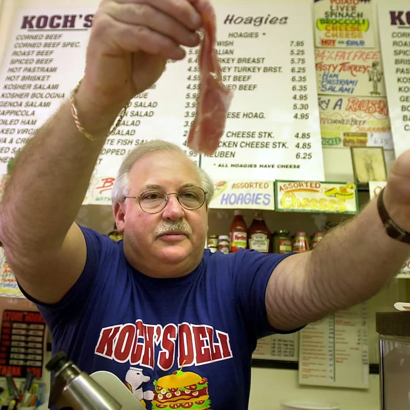A man in a Koch's Deli shirt holds up a slice of deli meat in front of a menu.