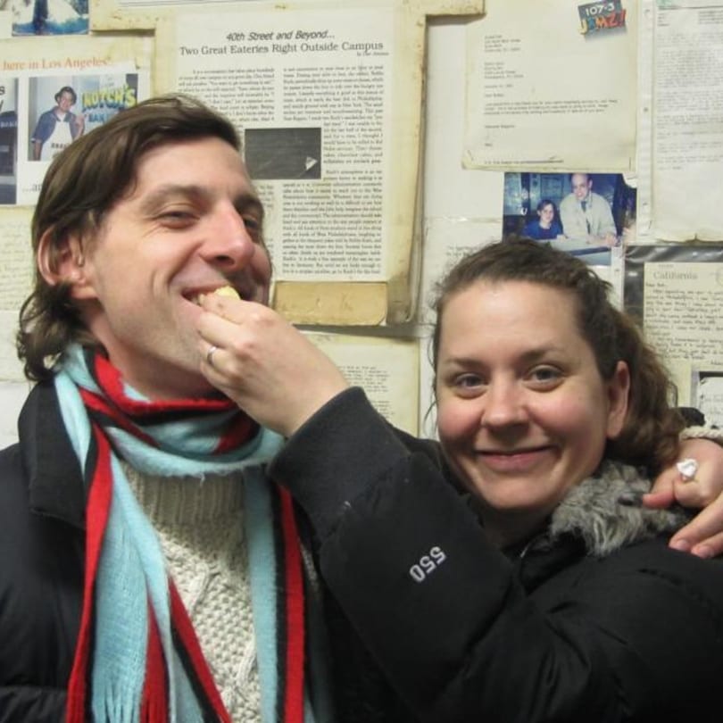 A woman feeds a smiling man a bite of food at Koch's Deli, with newspaper clippings in the background.